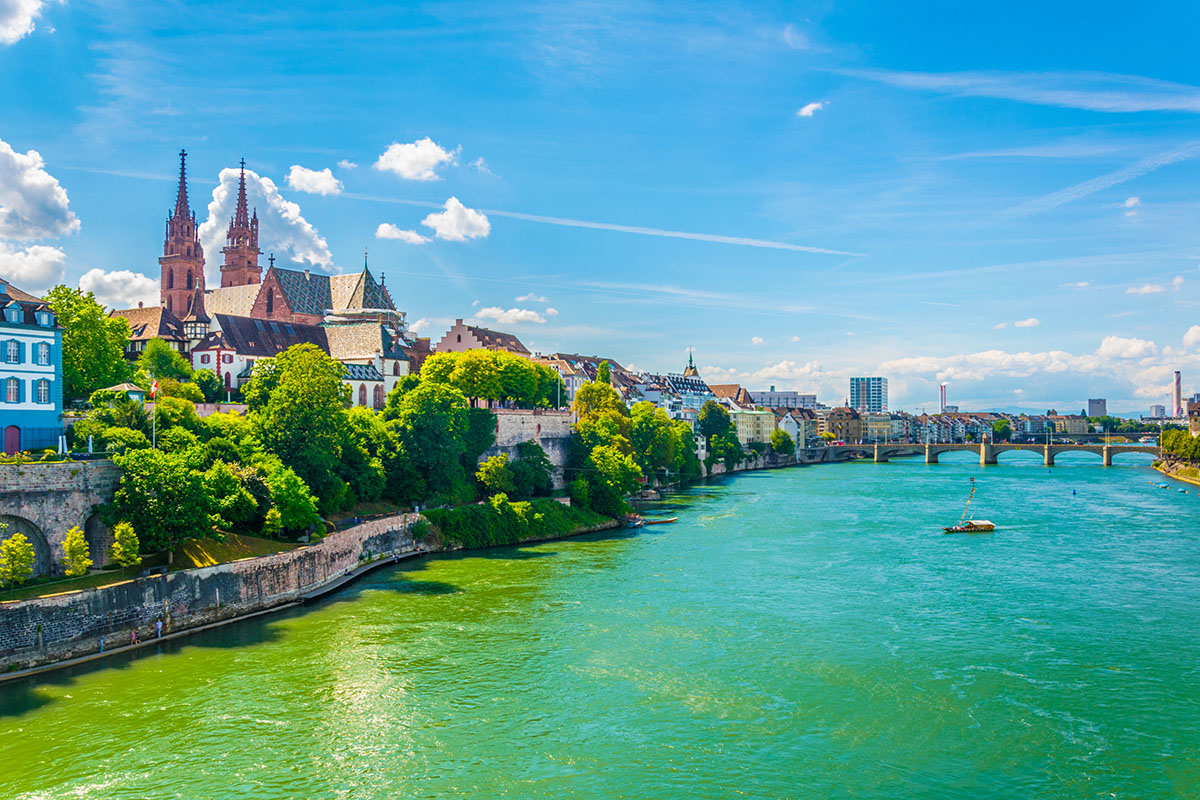 Basel, Switzerland A picturesque view of a river in Basel, Switzerland, featuring historic buildings with colorful facades, lush greenery, and the iconic twin spires of a cathedral against a bright blue sky with scattered clouds. A bridge spans the river, and a small boat navigates the tranquil waters.