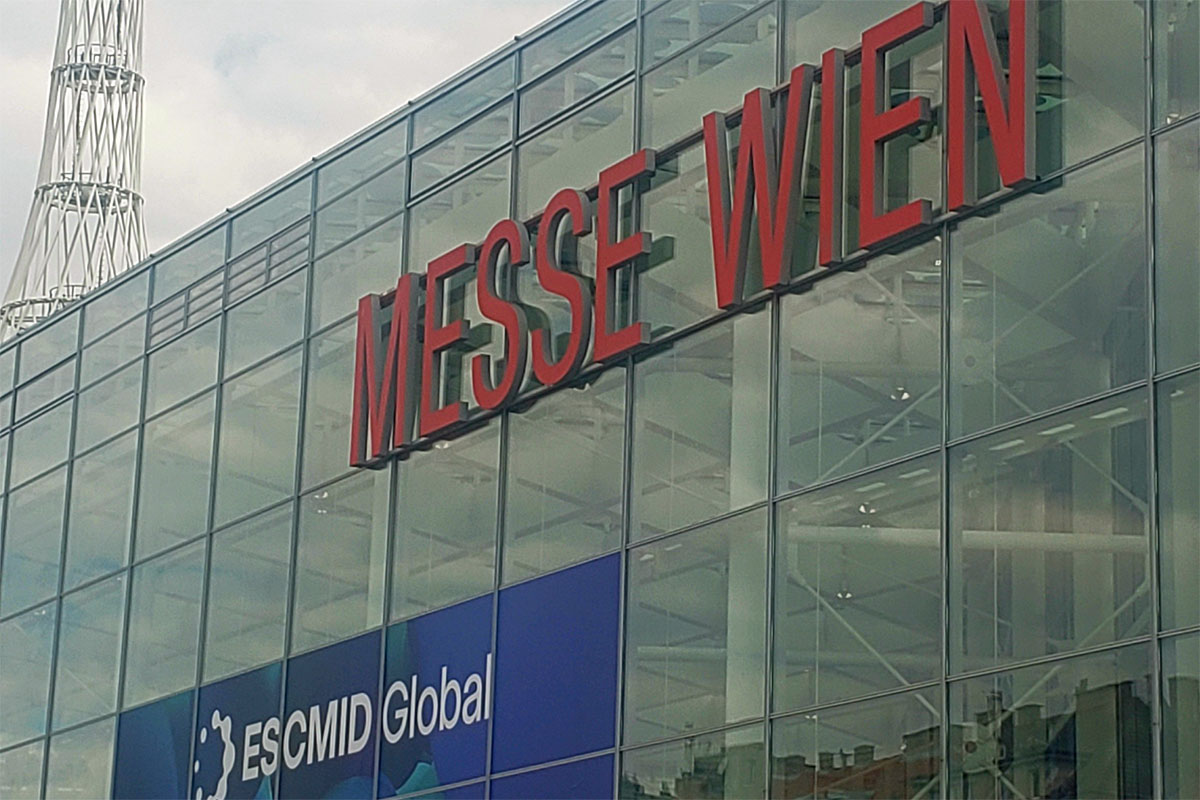 Exterior view of Messe Wien convention center featuring the "ESCMID Global" event banner, with people congregating at the entrance and a tall communication tower in the background.