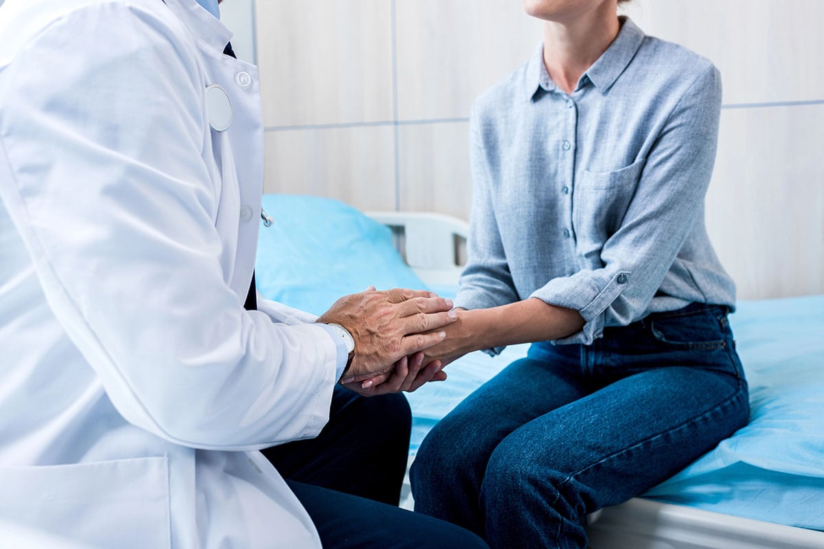 A healthcare professional in a white coat gently holds the hand of a patient sitting on a hospital bed, conveying a sense of care and empathy.